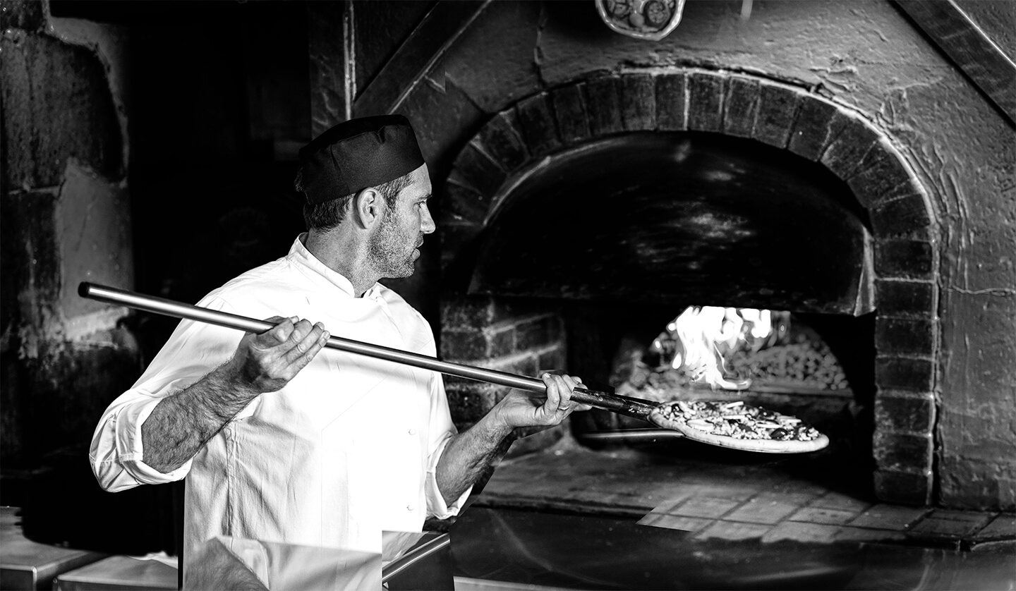 Un chef en uniforme blanc utilise une pelle à pizza pour enfourner une pizza dans un four à bois traditionnel